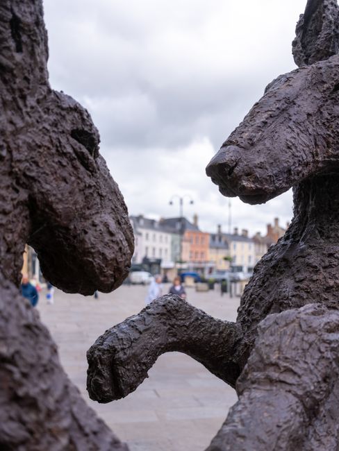 A close-up view of a sculpture featuring two large, textured bronze hands reaching out towards each other, set against a background of a town square with multicolored buildings, cobbled streets, and p